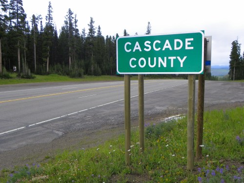 Sign for Cascade County beside a road, surrounded by trees and wildflowers.