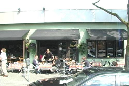 A bustling outdoor café with patrons dining at tables, surrounded by greenery and a sunny atmosphere.