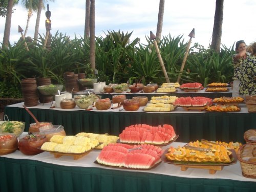 A vibrant outdoor buffet featuring a variety of fresh fruits, salads, and dishes, surrounded by tropical greenery.