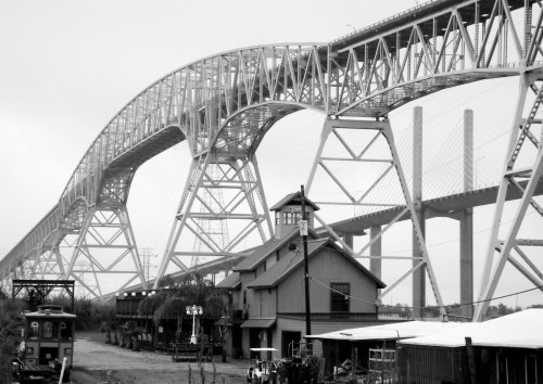 A black and white image of a large bridge arching over a rustic building and train tracks below.