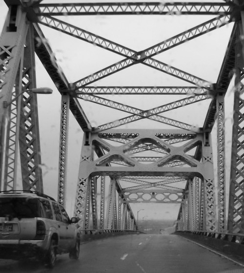 A black and white image of a metal bridge with intricate truss designs, viewed from a vehicle on a rainy day.