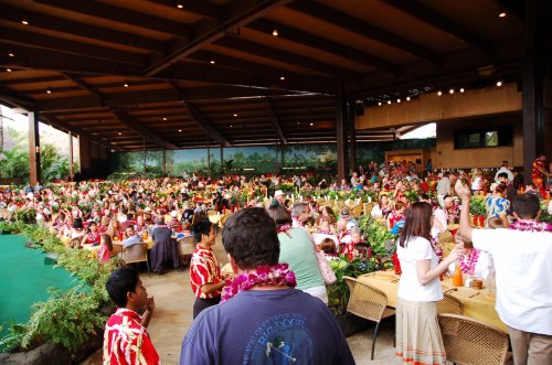 A large gathering of people in a tropical setting, enjoying a festive meal with tables decorated with flowers and greenery.