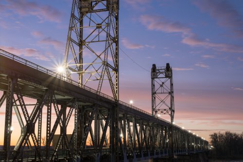 A metal bridge at sunset, with illuminated towers and a colorful sky in the background.