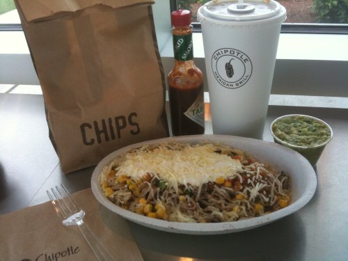 A Chipotle meal featuring a burrito bowl with cheese, chips, guacamole, and a drink on a table.