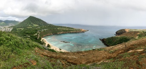 A panoramic view of a coastal landscape featuring a bay, turquoise waters, and lush green hills under a cloudy sky.