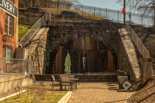 A stone archway under a road, with a small patio area featuring a table and umbrella in the foreground.