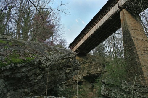 A covered bridge spans above rocky terrain, surrounded by trees and a clear blue sky.