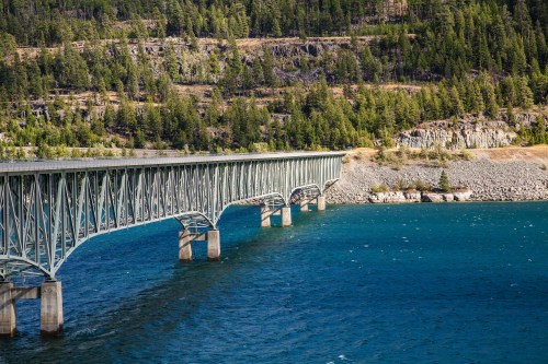 A long metal bridge spans over a blue river, surrounded by green trees and rocky cliffs.