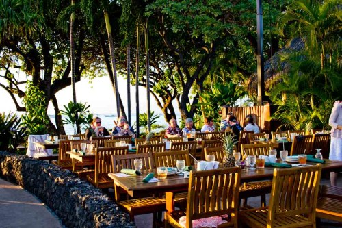 Outdoor dining area with wooden tables surrounded by lush greenery and palm trees, overlooking the ocean at sunset.