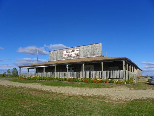 A wooden building with a sign reading "Ingalls Homestead," surrounded by flowers and a clear blue sky.