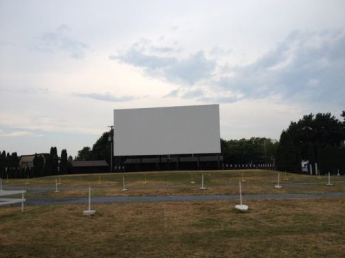 A large, blank outdoor movie screen stands on a grassy field under a cloudy sky, surrounded by white poles.