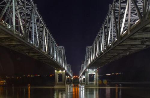 A lit steel bridge spans a river at night, reflecting lights on the water's surface.