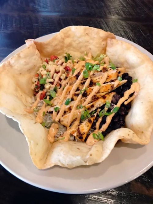 A tortilla bowl filled with rice, black beans, grilled chicken, and topped with sauce and green onions.