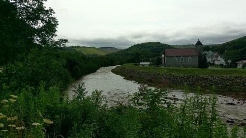 A serene river flows beside a church, with green hills and cloudy skies in the background. Wildflowers line the bank.