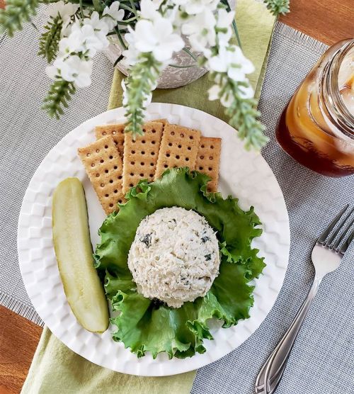 A plate with a rice dish on lettuce, a pickle, crackers, and a drink, with flowers in a vase in the background.