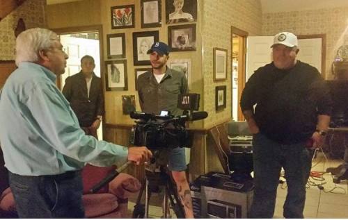 A man interviews two others in a cozy room filled with framed artwork and a video camera set up.