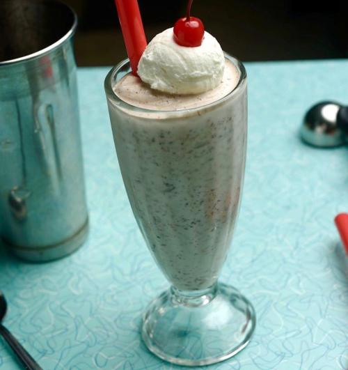 A tall glass of milkshake topped with whipped cream and a cherry, served with a red straw on a blue patterned table.