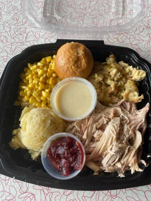 A takeout meal featuring shredded chicken, corn, stuffing, mashed potatoes, a roll, and two sauces in containers.