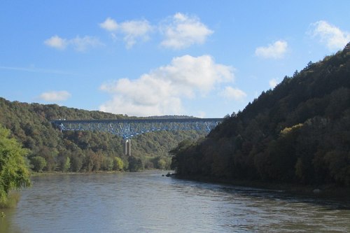 A blue bridge spans a river, surrounded by lush green hills under a partly cloudy sky.