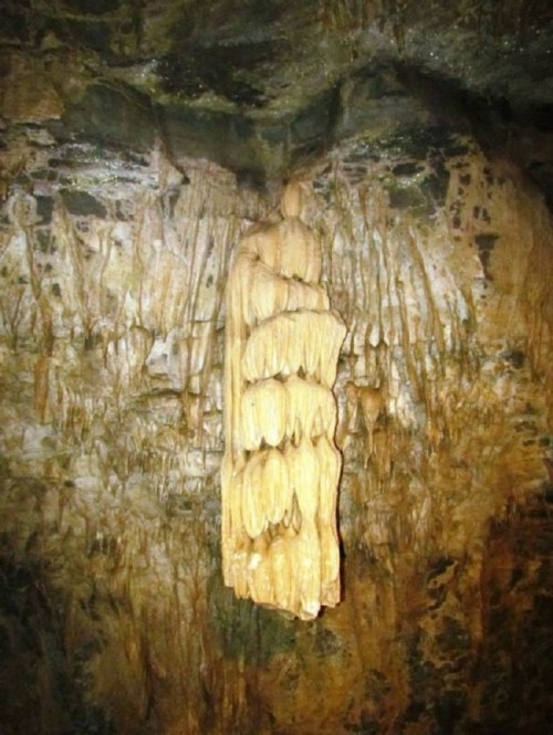 A large, elongated stalactite hangs from a rocky cave ceiling, surrounded by textured stone formations.