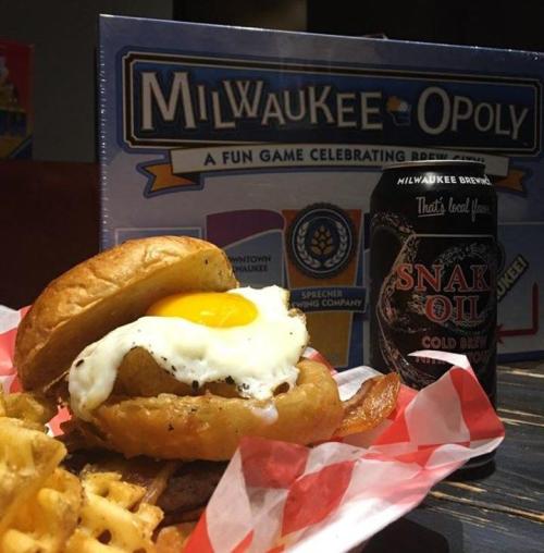 A burger with a fried egg, waffle fries, and a can of Snak Oil beer, with a Milwaukee-opoly game in the background.