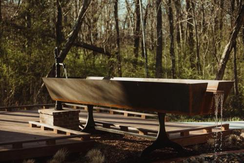 A rustic outdoor bathtub with a faucet, surrounded by trees and greenery on a wooden deck. Water flows from the faucet.