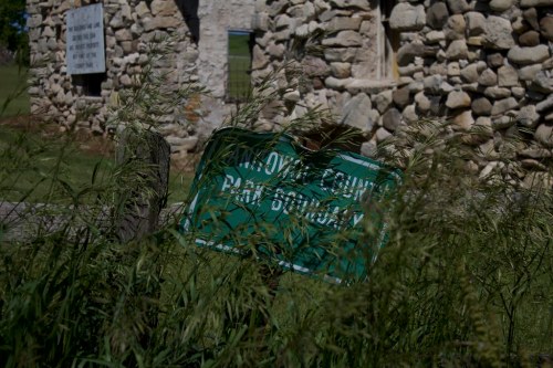 A weathered green sign partially obscured by tall grass, in front of a stone building.