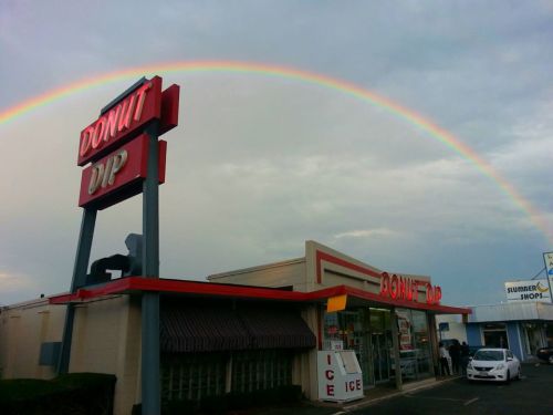 A colorful rainbow arcs over the Donut Dip shop, showcasing its retro sign and inviting storefront.