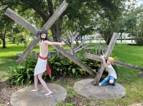 A sculpture of a man in a robe and a child, both interacting with large wooden crosses in a green park setting.