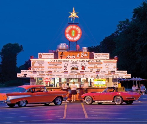 A retro drive-in diner with neon lights, classic cars parked outside, and a menu board filled with vintage food options.