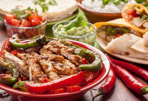 A vibrant spread of grilled chicken with peppers, tortillas, rice, guacamole, and fresh salsa on a wooden table.