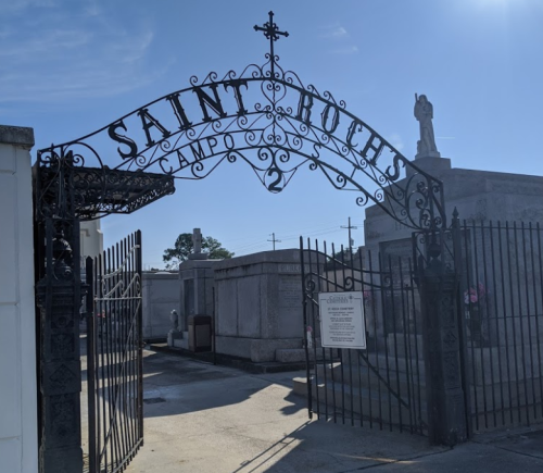 Wrought iron gate of Saint Roch Cemetery, featuring a cross and angel statue, under a clear blue sky.