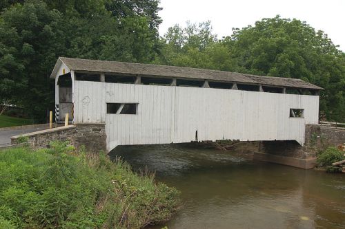 A white, weathered covered bridge spans a calm stream, surrounded by lush greenery and trees.