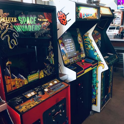Three vintage arcade machines lined up, featuring "Deluxe Space Invaders" and colorful designs.