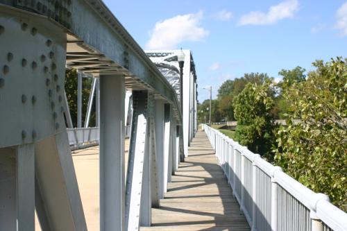 A metal bridge with wooden planks, lined with trees, under a clear blue sky.