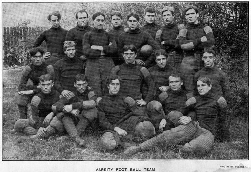 A vintage photo of a varsity football team, featuring 15 players in dark uniforms, posed outdoors.