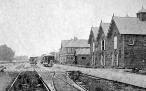 Historic black-and-white photo of a streetcar line with wooden buildings and tracks in a rural setting.