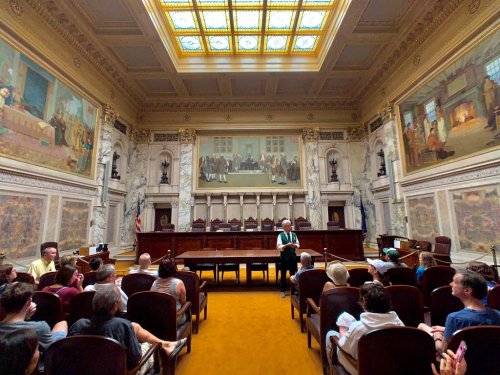 A historic courtroom with ornate decor, featuring a speaker and an audience seated in wooden chairs.