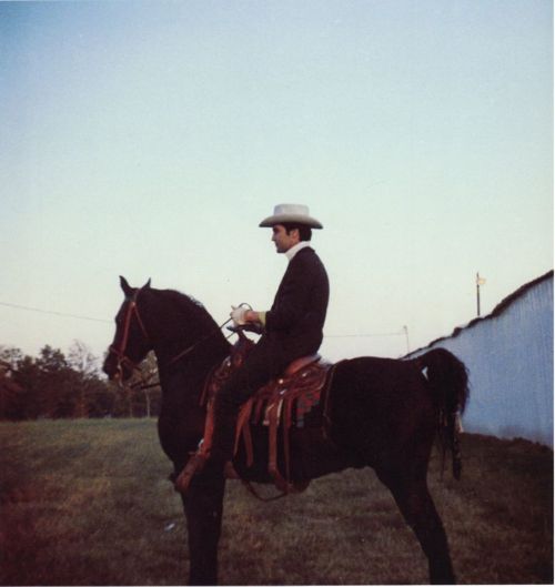 A man in a cowboy hat sits on a black horse in an open field, holding something in his hands.