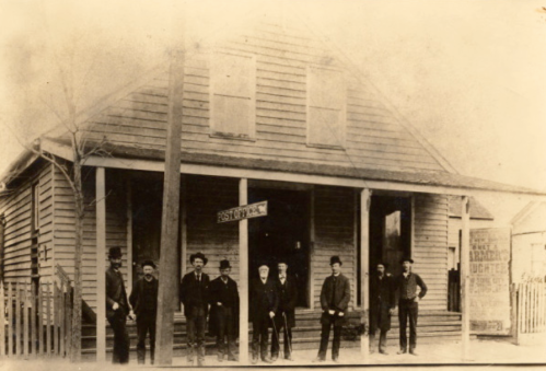 A historic black-and-white photo of a group of men standing outside a wooden building with a sign reading "Post Office."