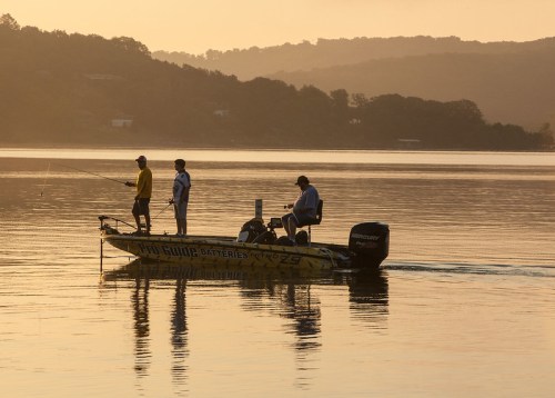 Two fishermen on a yellow boat casting lines into a calm lake at sunrise, surrounded by misty hills.
