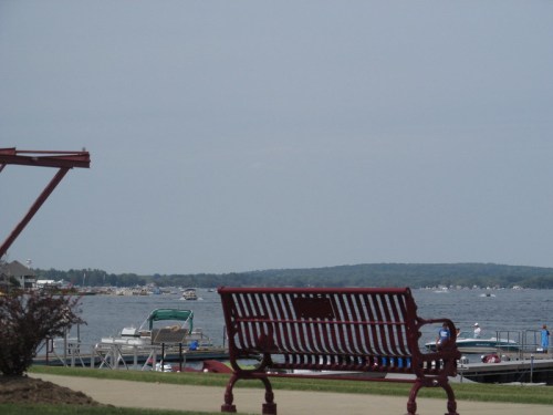 A red bench overlooks a calm lake with boats and a distant shoreline under a cloudy sky.