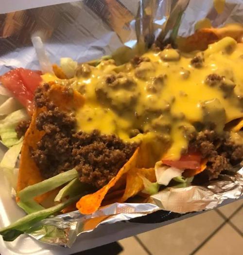 A close-up of a loaded nacho dish with ground beef, melted cheese, lettuce, and tomatoes in a foil container.
