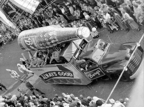 A vintage truck with a large soda bottle on top, surrounded by a crowd at a festive event.