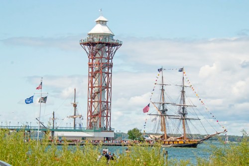 A tall lighthouse beside a tall ship on a calm waterway, with flags flying and greenery in the foreground.