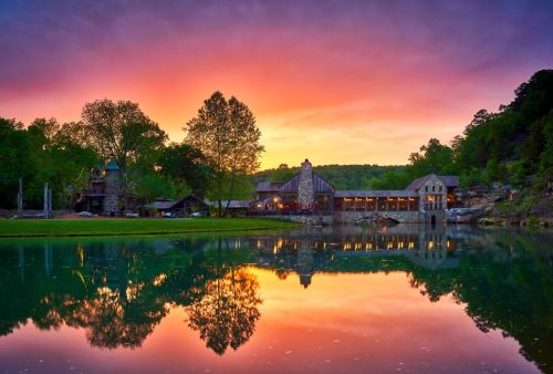 A serene sunset over a lake, reflecting a rustic building and lush trees against a colorful sky.