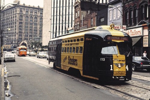 A vintage streetcar in black and yellow, parked on a city street with buildings and cars in the background.