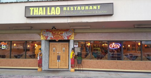 Exterior of Thai Lao Restaurant featuring a sign, open neon light, and decorative statues at the entrance.