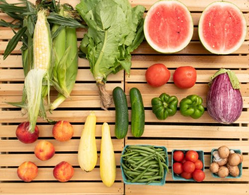 A variety of fresh vegetables and fruits arranged on a wooden surface, including corn, lettuce, watermelon, and tomatoes.