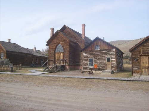 Historic wooden buildings in a ghost town, featuring a large church and smaller houses, set against a dry landscape.